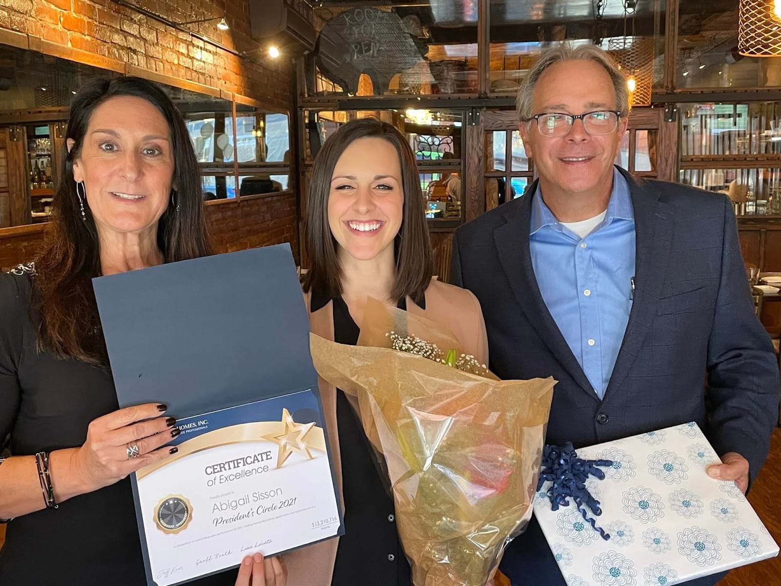 two women and man holding up certificate, flowers, and gift