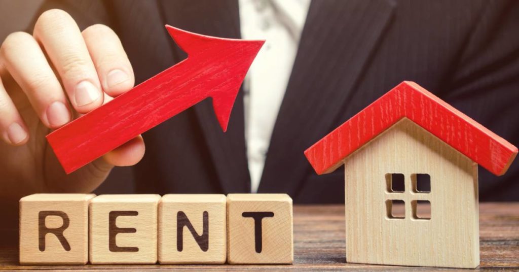 stock photo of rent spelled out in blocks next to a house with a hand holding an arrow going up